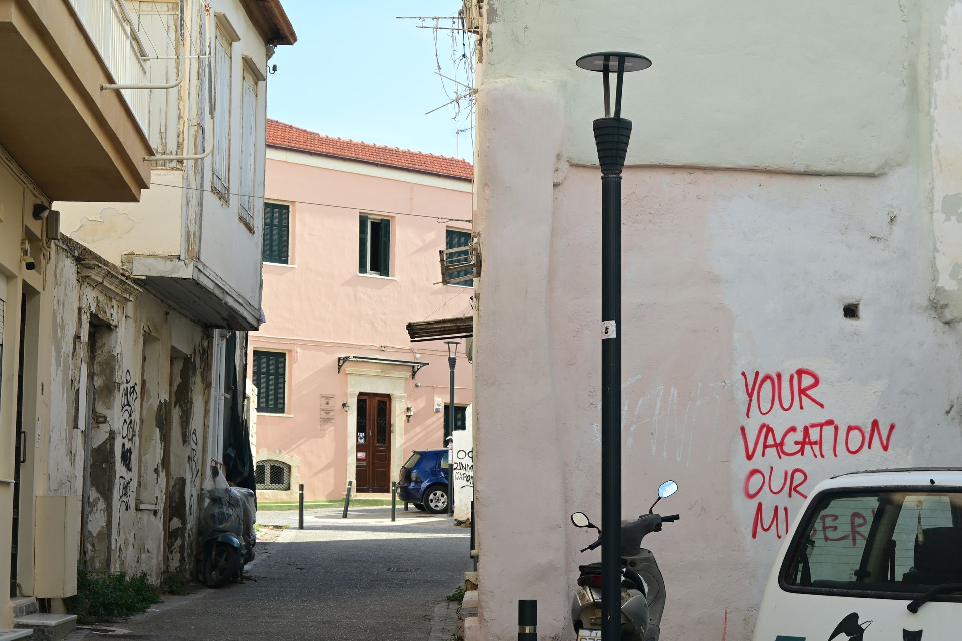 Narrow street with worn facades and graffiti reading Your vacation our misery