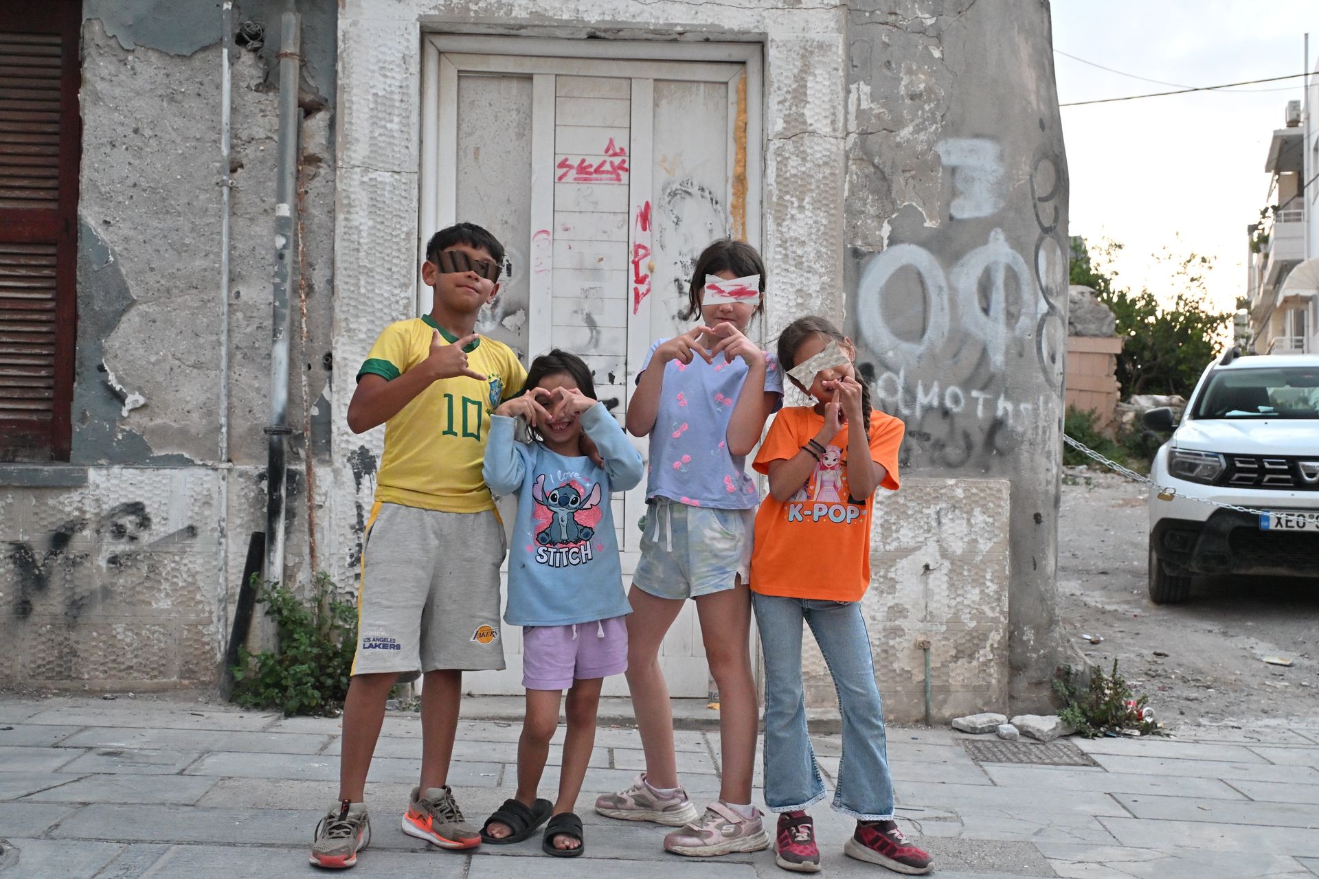 Four children standing outside a weathered building on a street with a parked car
